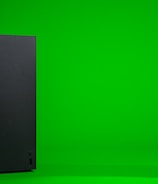A black, rectangular gaming console stands upright against a vibrant green background. The console has a simple, sleek design with minimalistic features including a disc slot and a few buttons.